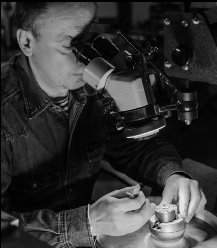 A black and white photograph of a watchmaker using a microscope to examine a small watch component on a workbench.